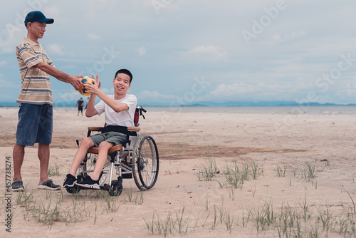 Happy disabled teenage boy on wheelchair playing a ball, Activity outdoors with father on the beach background, People having fun and diverse people concept.