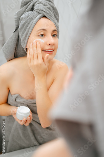 Portrait of a woman with a towel on her head in front of a mirror smearing white cream on her face. Concept of body care.