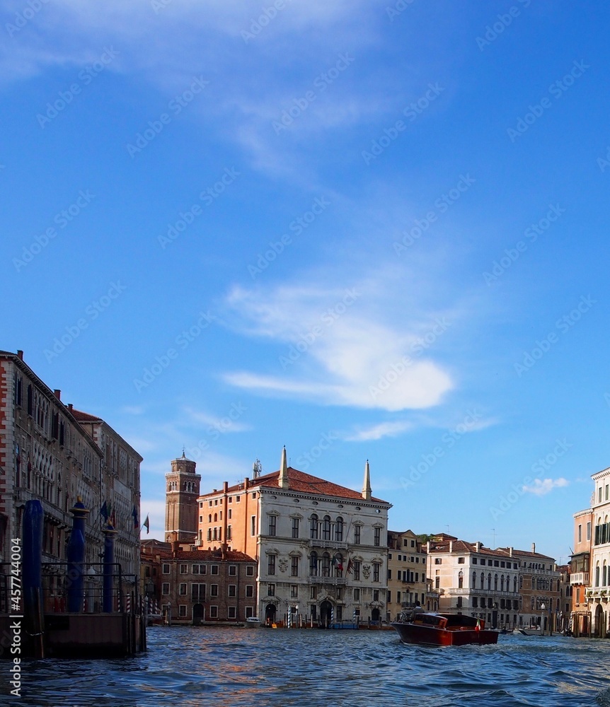 Venice, Italy - Grand Canal with blue sky and wispy clouds.  Palaces line the banks of the canal, and a speedboat navigates the center of the waterway. Vertical with modern colors.