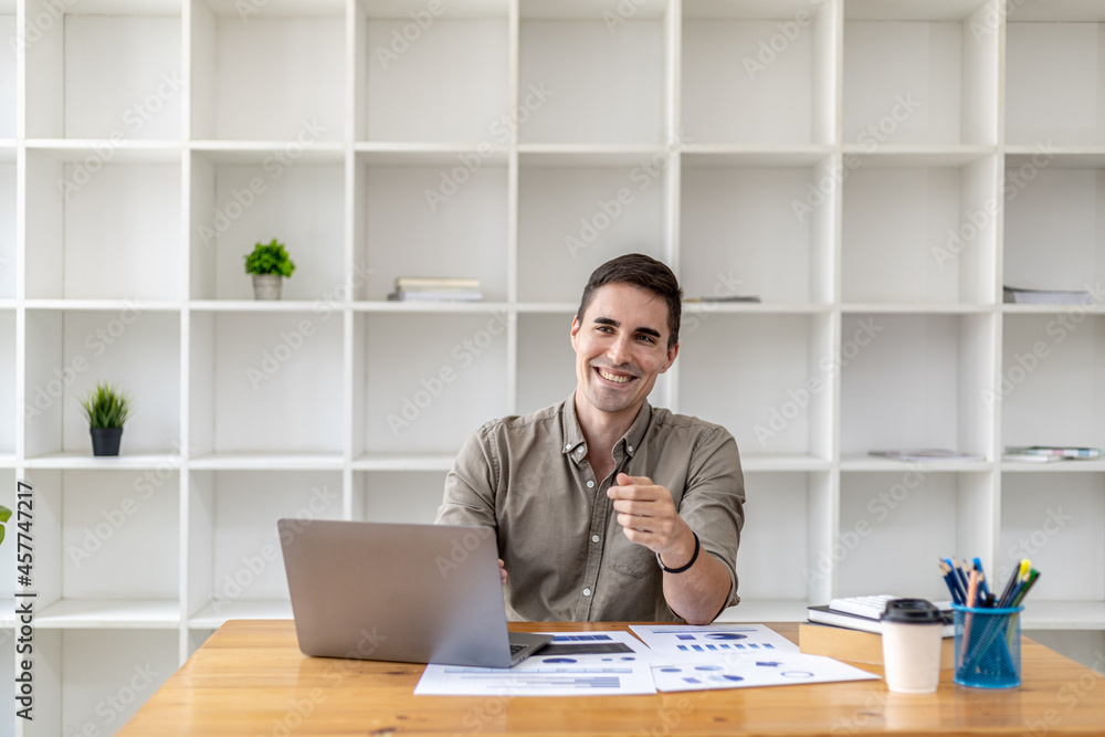 Fototapeta premium Young businessman sitting in the office working with laptop and paperwork on the desk, smiling, he is the founder of a startup, he is a young businessman who is skilled in running a growing company.