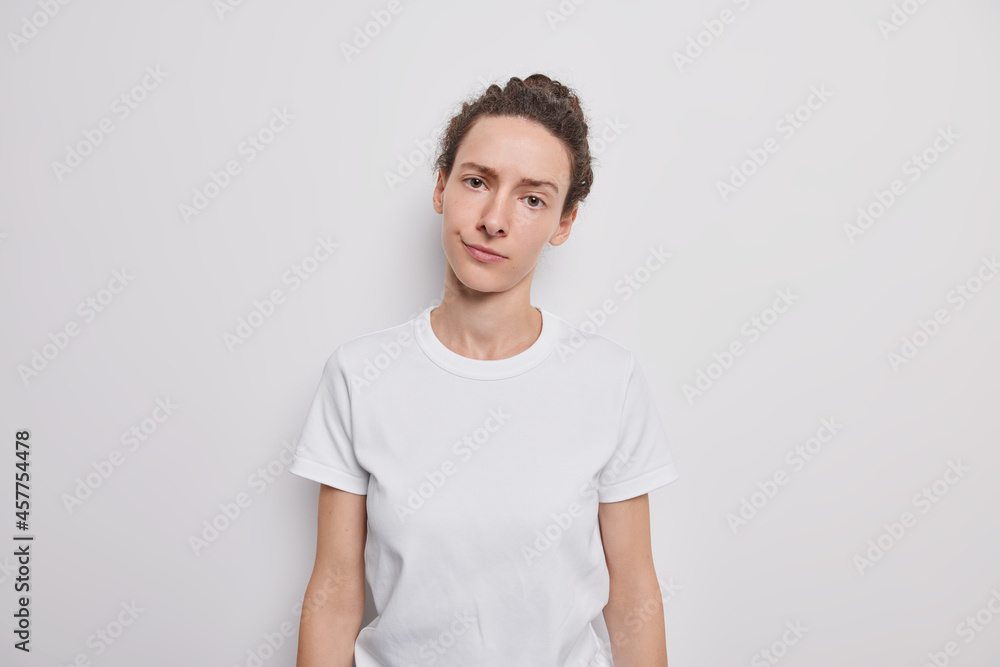 Studio shot of serious dark haired woman looks directly at camera purses lips looks directly at camera wears casual t shirt tilts head poses against white background has desperate need. Attentive gaze Studio shot of serious dark haired woman looks directly at camera purses lips looks directly at camera wears casual t shirt tilts head poses against white background has desperate need. Attentive gaze
