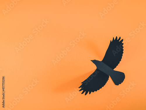 Black paper crow on an orange background. © Nataliya