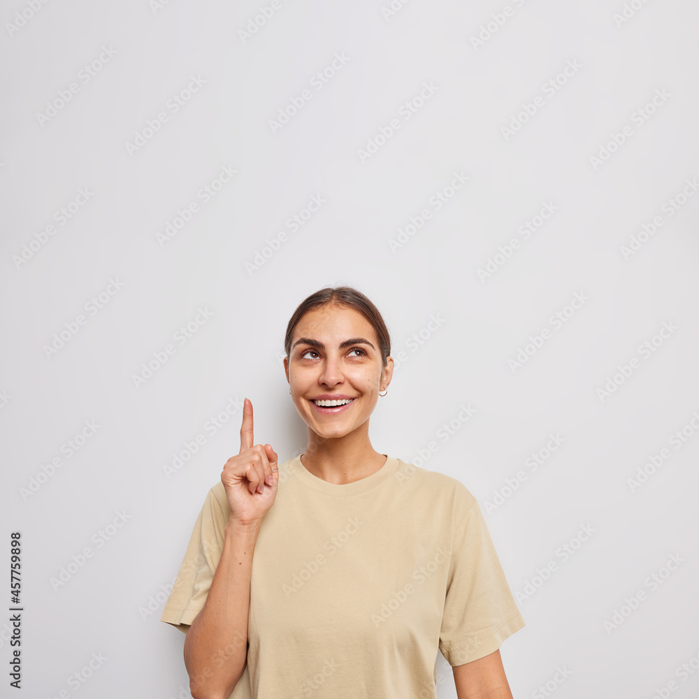 Vertical shot of positive European woman points above with index finger shows advertisement or promo offer dressed in casual t shirt isolated over white background chooses smth in store. Hey see this. Vertical shot of positive European woman points above with index finger shows advertisement or promo offer dressed in casual t shirt isolated over white background chooses smth in store. Hey see this.