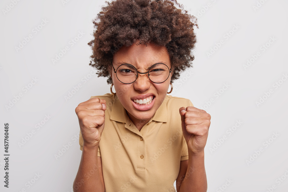 Afro American woman clenches teeth and fists angrily being irritated with something cannot stand annoying noise wears spectacles casual t shirt isolated over grey background. Negative emotions - obrazy, fototapety, plakaty 