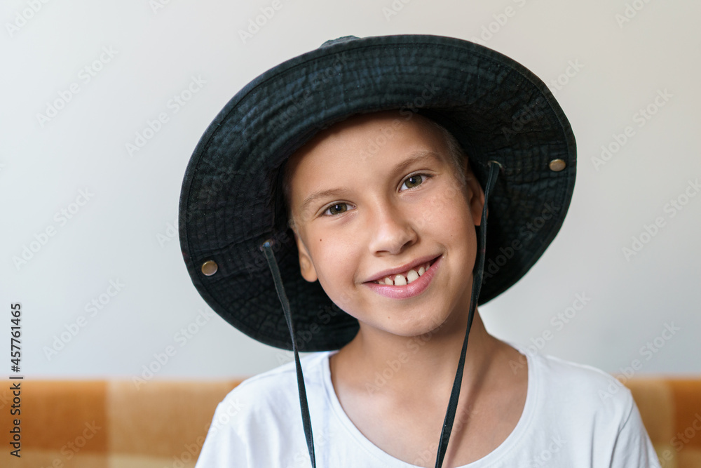 Cheerful school-age boy smiling while looking at the camera. In a white t-shirt and a black sun hat. Caucasian child having fun at the camera while sitting at home. Cheerful school-age boy smiling while looking at the camera. In a white t-shirt and a black sun hat. Caucasian child having fun at the camera while sitting at home.