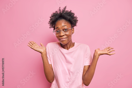 Young dark skinned woman shrugs shoulders shows empty hands has no clue about something nothing to say stands unaware indoor wears big optical glasses casual t shirt isolated over pink wall.