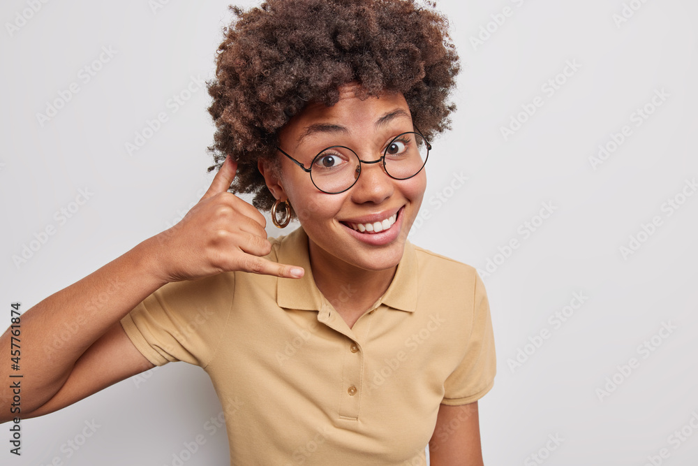 Positive African American woman with curly hair shows mobile phone gesture near ear tells to contact her says call me smiles happily wears round spectacles casual brown t shirt poses indoor. Positive African American woman with curly hair shows mobile phone gesture near ear tells to contact her says call me smiles happily wears round spectacles casual brown t shirt poses indoor.