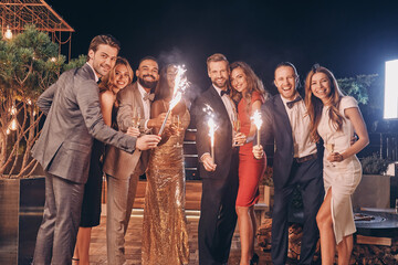 Group of beautiful people in formalwear holding sparklers and smiling while spending time on outdoor party