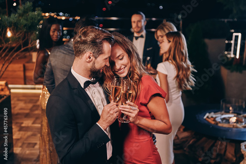 Beautiful young couple in formalwear holding champagne flutes and smiling while spending time on luxury party photo