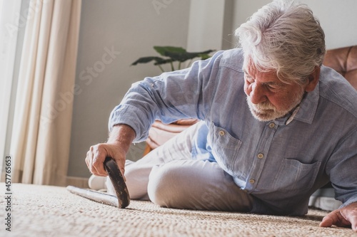 Elder senior man lying on floor after falling down with wooden walking stick beside couch on rug in living room at home. Old man suffering with pain and struggling to get up after falling down at home photo