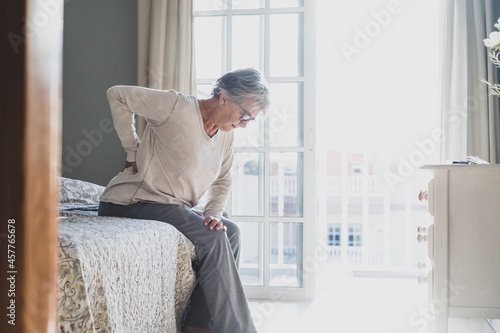 Senior woman suffering from backache after sleep, rubbing stiff muscles, Old female sitting on bed touching lower back feeling discomfort because of uncomfortable bed at home