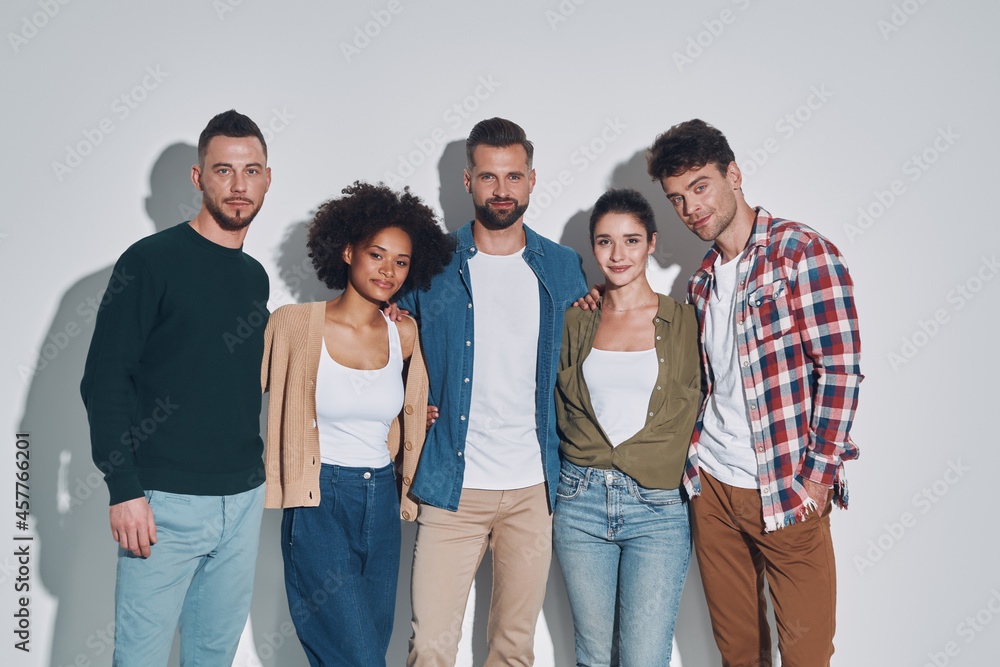 Group of young beautiful people in casual clothing bonding and smiling while standing against gray background