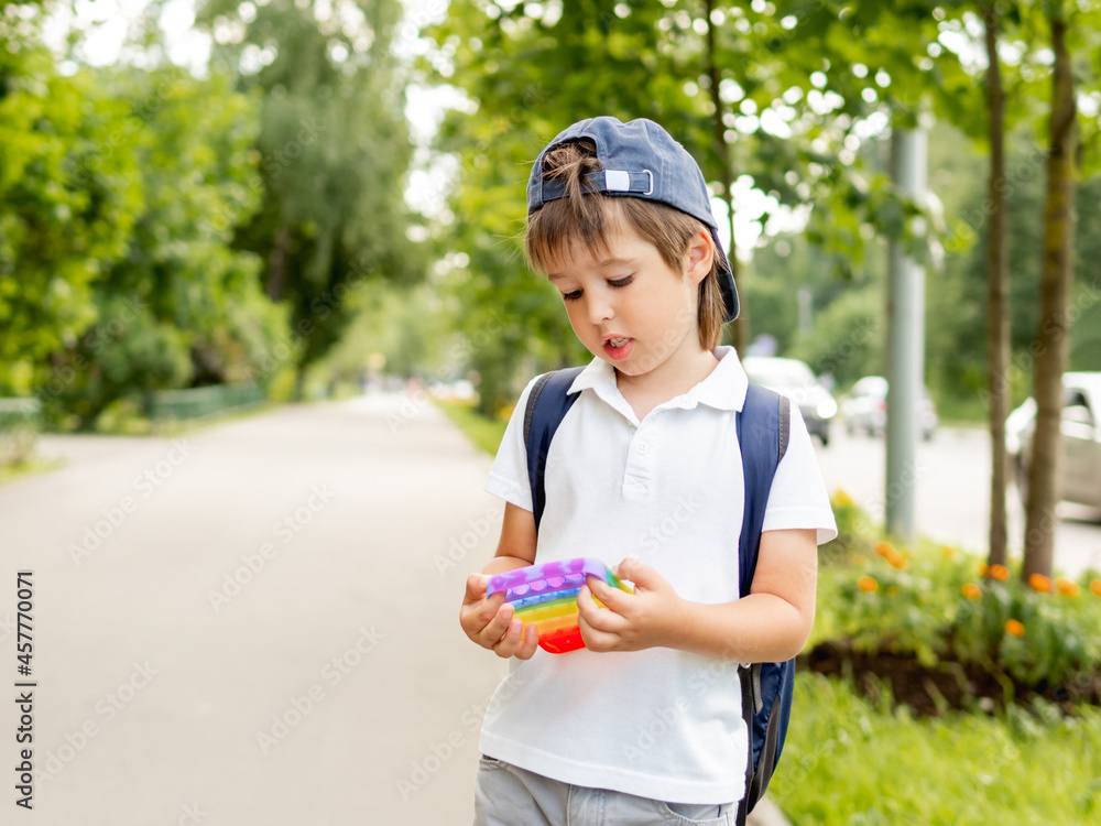 Little boy plays square rubber fidget toy Pop It. Rainbow-colored anti stress and tactile toy for all ages. Outdoor recreation with toy for fine motor skills improvement. Little boy plays square rubber fidget toy Pop It. Rainbow-colored anti stress and tactile toy for all ages. Outdoor recreation with toy for fine motor skills improvement.