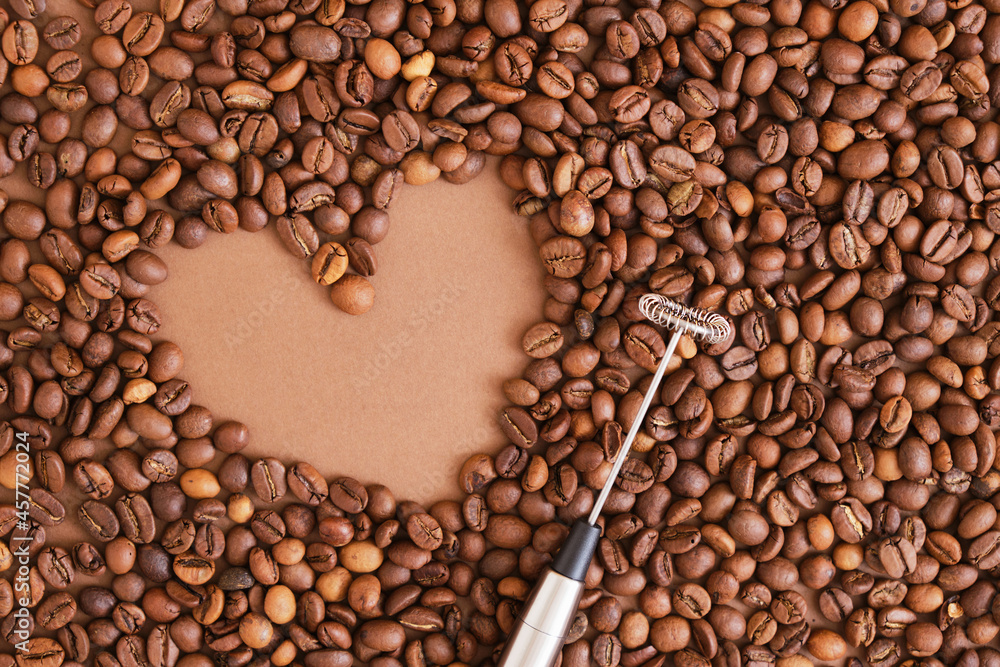 heart made of coffee beans and metal handheld milk steamer on brown background. Handheld frother heart made of coffee beans and metal handheld milk steamer on brown background. Handheld frother