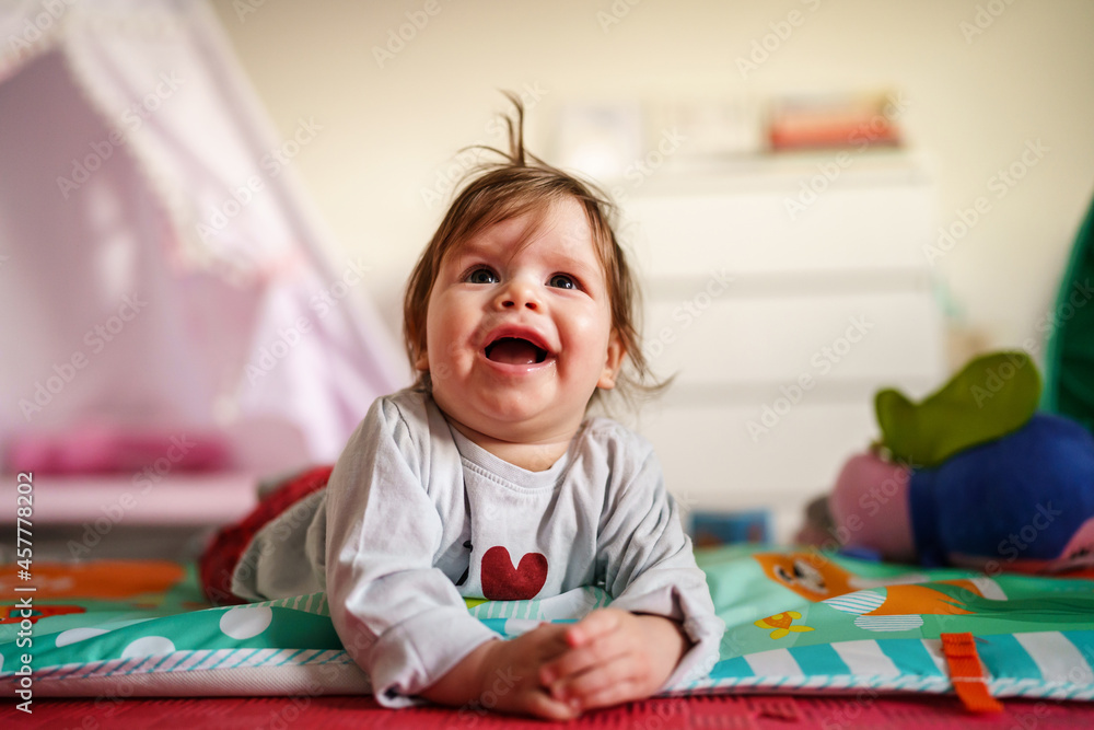 Obraz premium Baby small caucasian girl five months old lying belly down on tummy at home in summer day playing with toy childhood concept looking to the camera alone front view smiling
