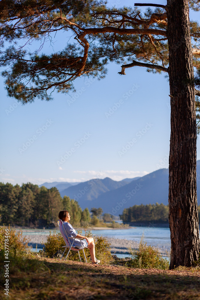 A woman is sitting on a folding chair on the bank of a mountain river on a nice, warm day under a large tree. A calm and quiet place to relax and reflect. Equipment and a tourist's rest. A woman is sitting on a folding chair on the bank of a mountain river on a nice, warm day under a large tree. A calm and quiet place to relax and reflect. Equipment and a tourist's rest.