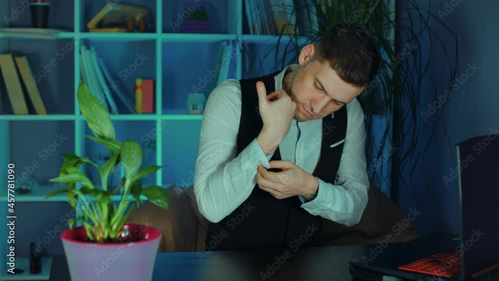 Young man fastens buttons on his sleeves, sitting in armchair before computer in office. Business employee preparing for work on laptop. Young man fastens buttons on his sleeves, sitting in armchair before computer in office. Business employee preparing for work on laptop.