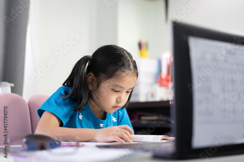 Asian child student or kid girl reading on book to learning and studying online class on computer notebook or person learn from home by video call on laptop for back to school by homeschool education