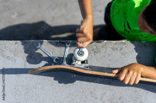 Top view of a concentrated child repairing his skateboard before using it inside the skateboard park on a sunny day. High quality photo