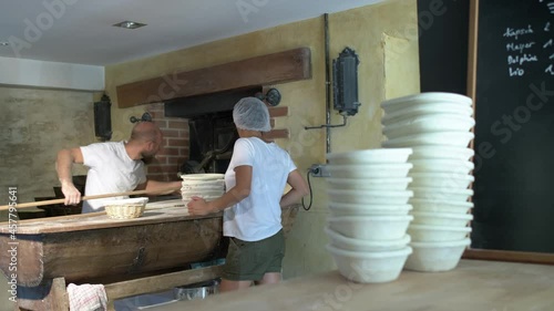 Large shot, background, a baker flouring, cuting and puting bread loaf in a traditional stone oven, using baker’s peel, foreground a women is managin bread’s baskets photo