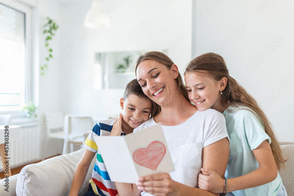 Joyful young mother and little children sitting on sofa and embracing while reading wishes and congratulations with holiday in presented postcard at home Joyful young mother and little children sitting on sofa and embracing while reading wishes and congratulations with holiday in presented postcard at home