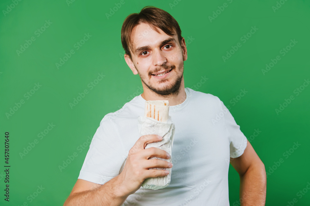 A young man in a white T-shirt on a green background holds lunch, a doner with chicken. A young man in a white T-shirt on a green background holds lunch, a doner with chicken.