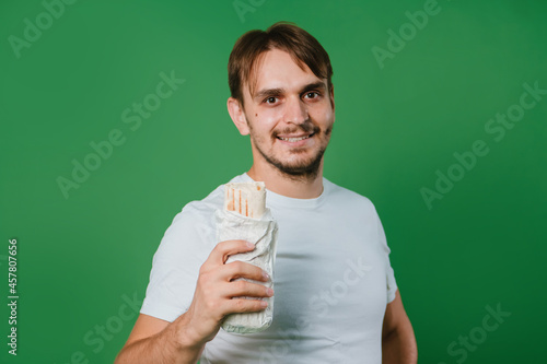 A young man in a white T-shirt on a green background holds lunch, a doner with chicken.