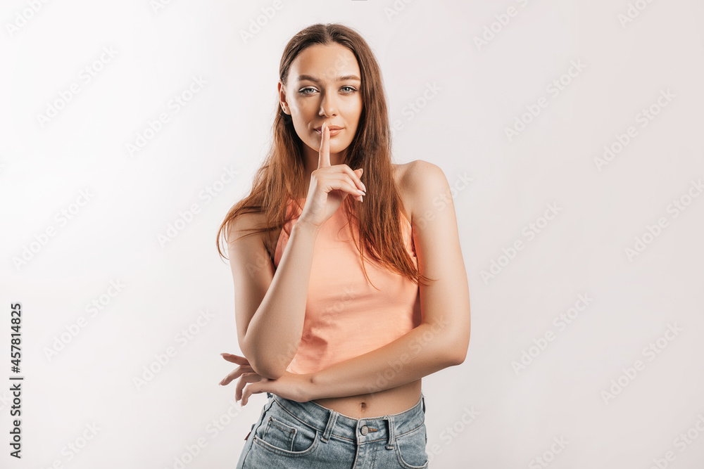 Shh gesture. Young beautiful serious girl holding a finger to her lips on a white isolated background. A woman asks to be silent, a place for advertising. Positive brunette in orange top Shh gesture. Young beautiful serious girl holding a finger to her lips on a white isolated background. A woman asks to be silent, a place for advertising. Positive brunette in orange top
