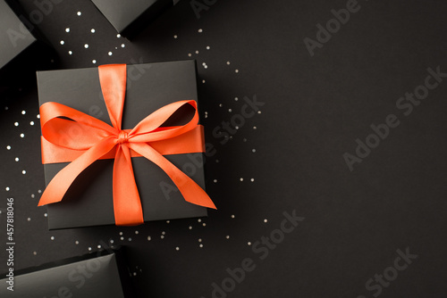 Top view photo of stylish black gift boxes with orange ribbon bow and sequins on isolated black background with copyspace