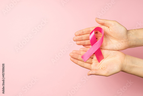 First person top view photo of young girl's hands holding pink satin ribbon in palms symbol of breast cancer awareness on isolated pastel pink background with empty space