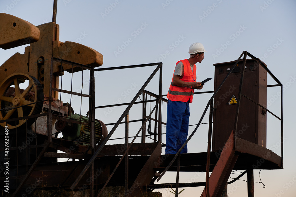 Male worker in protective helmet holding clipboard while monitoring work of petroleum pump jack. Petroleum engineer in work vest working in oil field. Concept of petroleum industry and oil extraction. Male worker in protective helmet holding clipboard while monitoring work of petroleum pump jack. Petroleum engineer in work vest working in oil field. Concept of petroleum industry and oil extraction.