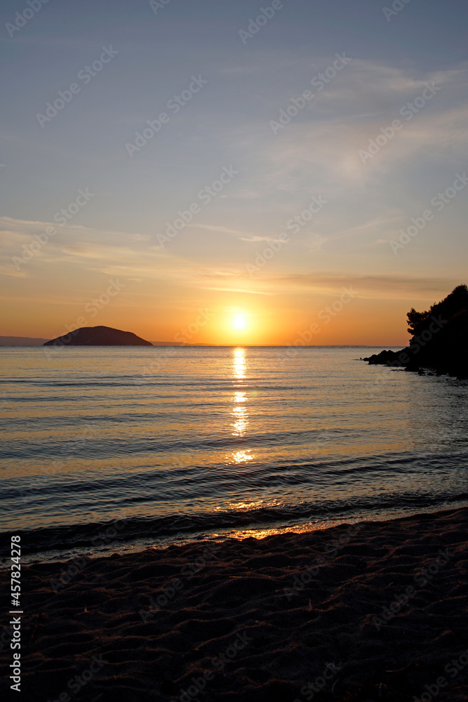Bright red sunset over the Aegean Sea. Sun and mountains. Horizon. Waves and glare. Beach. Vertical shot