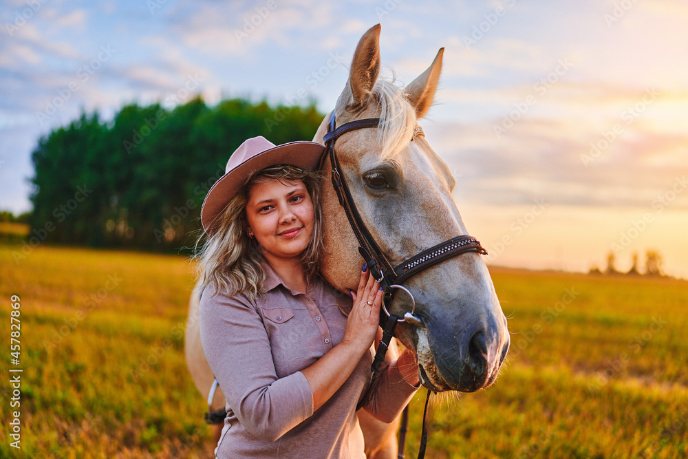 Fototapeta premium Portrait of young cute happy joyful satisfied smiling woman hugging and stroking beautiful palomino blond horse at meadow at sunset