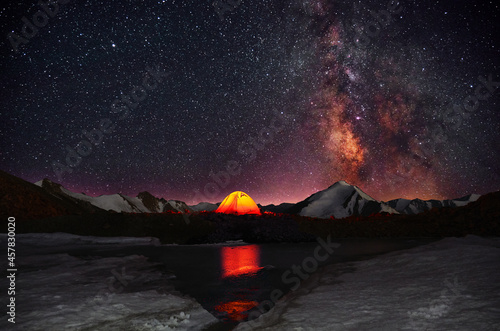 Brightly glowing orange tent set up in a romantic place on the shore of a glacial lake on the background of snow-capped mountain peaks and a starry sky with the Milky Way © kiwisoul
