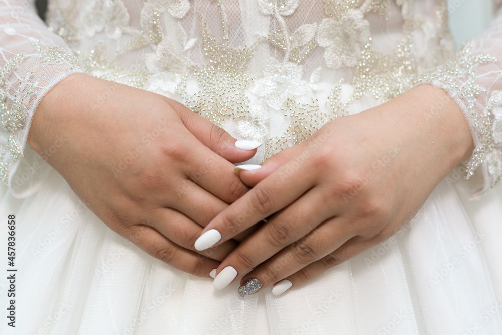 Delicate hands of the bride with a white manicure on the background of the wedding dress.