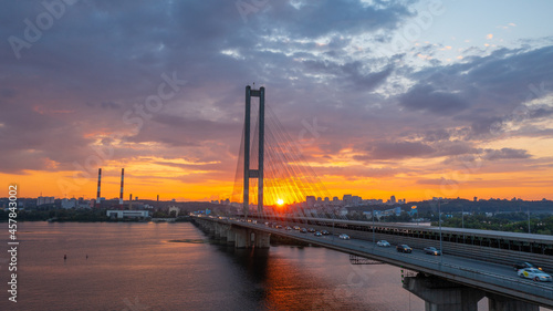 South bridge in Kiev. Sunset over the Dnieper. Thick clouds over the evening city. Evening shot of the bridge. Orange sun at sunset. The rays break through the clouds and are reflected in the river.