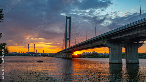 South bridge in Kiev. Sunset over the Dnieper. Thick clouds over the evening city. Evening shot of the bridge. Orange sun at sunset. The rays break through the clouds and are reflected in the river.