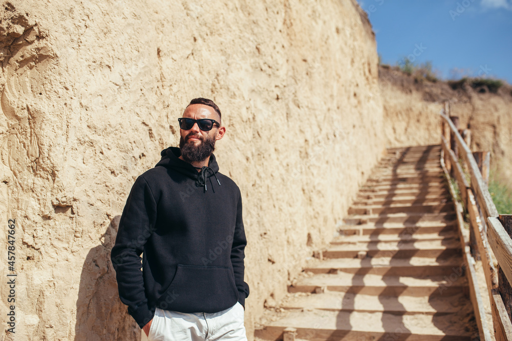 Young bearded hipster guy with beard in black blank hoodie or sweatshirt and sunglasses at the tropical beach. Mock-up for print.