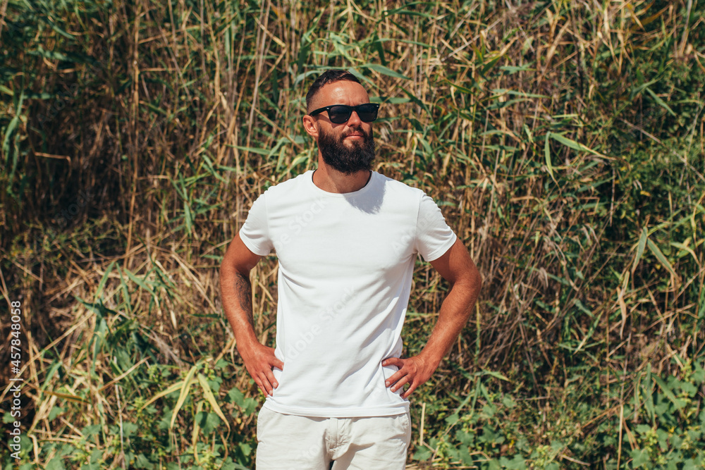 Fototapeta premium Young bearded hipster guy with beard in white t-shirt and sunglasses at the tropical beach. Mock-up for print. T-shirt template.