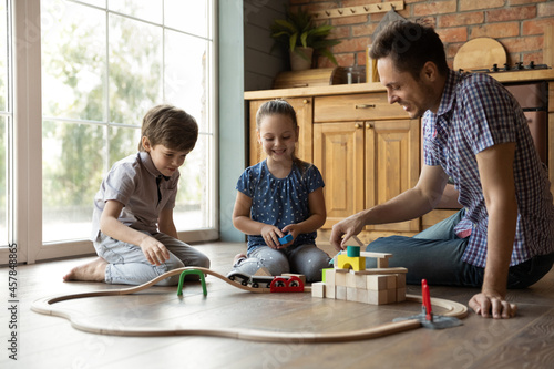 Joyful adorable little kids brother sister playing with toy railway road, sitting on heated warm wooden floor with caring smiling young father in kitchen, enjoying carefree weekend leisure pastime.