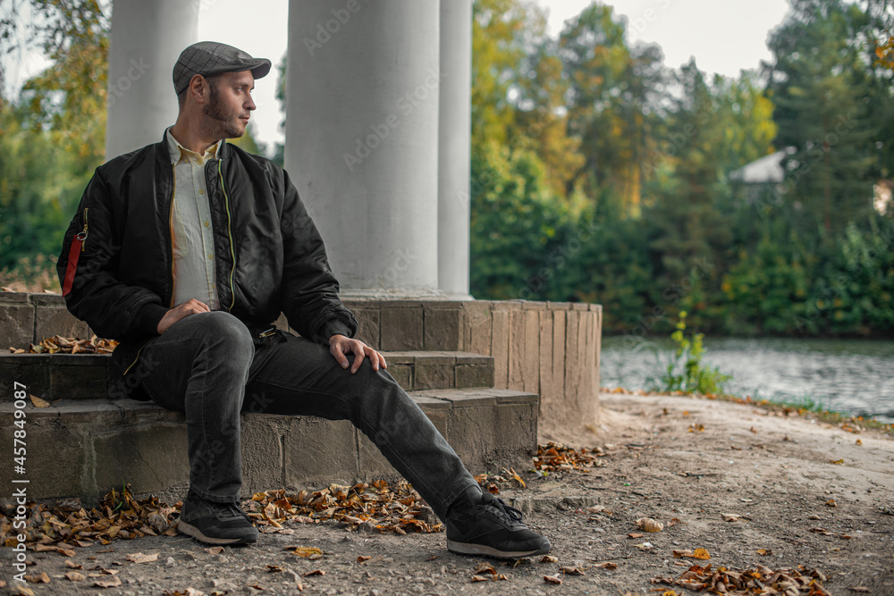 Fototapeta premium a serious man in black sits on the steps in the autumn by the river in a park