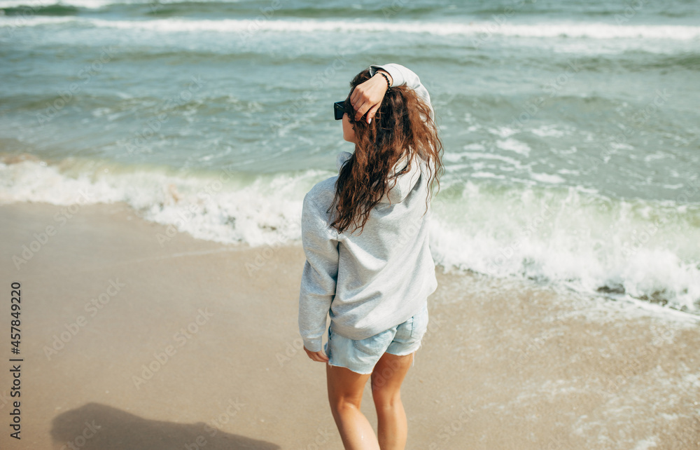Beautiful young adult woman in gray blank hoodie or sweatshirt and sunglasses at the tropical beach. Mock-up for print. Space for your logo or design. Beautiful young adult woman in gray blank hoodie or sweatshirt and sunglasses at the tropical beach. Mock-up for print. Space for your logo or design.