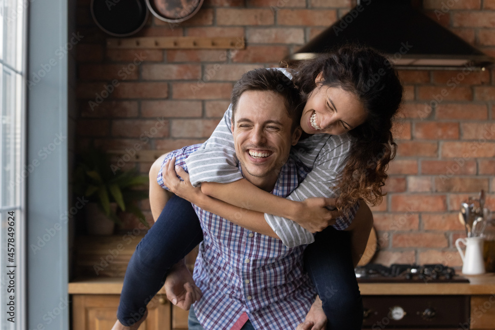 Happy young latin man giving piggyback ride to laughing attractive wife, having fun together in own cottage house, celebrating moving day. Cheerful millennial family couple entertaining on weekend.