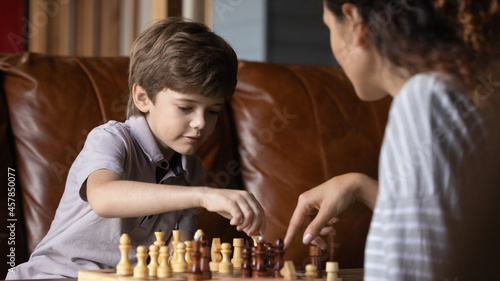 Fotografie Concentrated small adorable child boy learning playing chess with caring young mother or babysitter, planning next moves, developing strategic skills, improving memory, enjoying hobby activity
