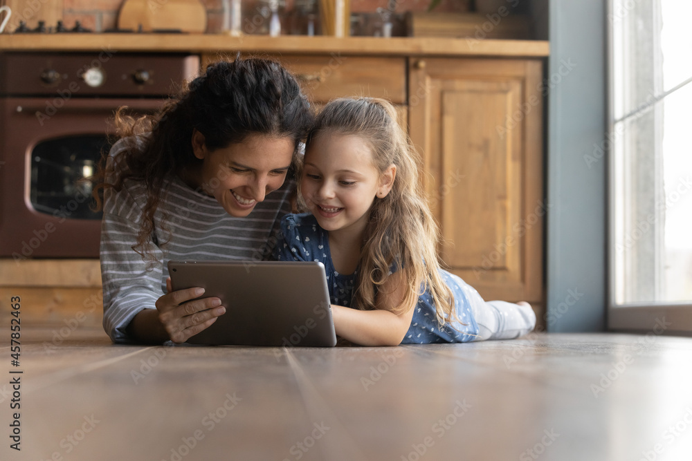 Happy young mother watching cartoons or funny photo video content in social networks on tablet with cute smiling little kid daughter, lying together on heated warm floor, tech addiction concept. Happy young mother watching cartoons or funny photo video content in social networks on tablet with cute smiling little kid daughter, lying together on heated warm floor, tech addiction concept.