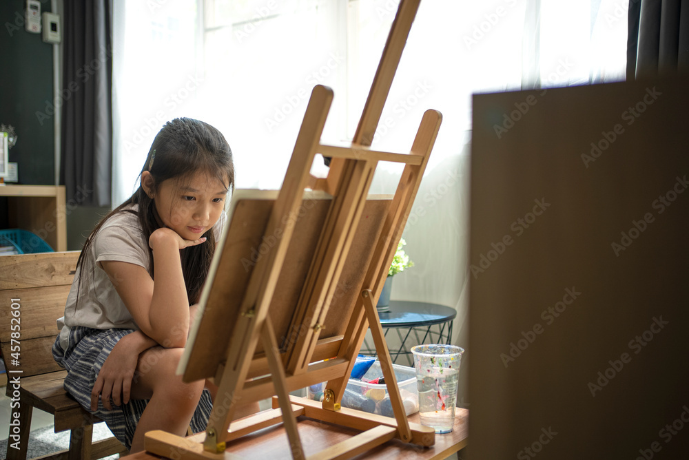 Asian beautiful girl with brush nd palette painting water color to canvas on wooden easel at home for her relaxing time.