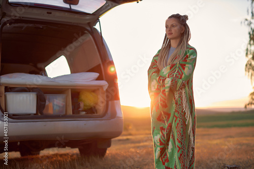 a bright young woman stands in a field at sunset near her car and looks into the distance. High quality photo