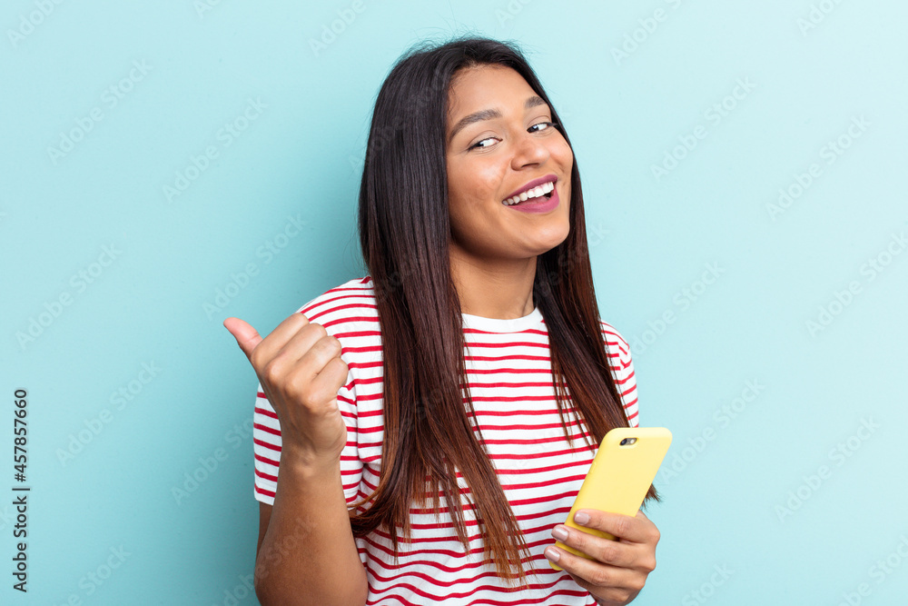 Young Venezuelan woman holding mobile phone isolated on blue background points with thumb finger away, laughing and carefree. Young Venezuelan woman holding mobile phone isolated on blue background points with thumb finger away, laughing and carefree.