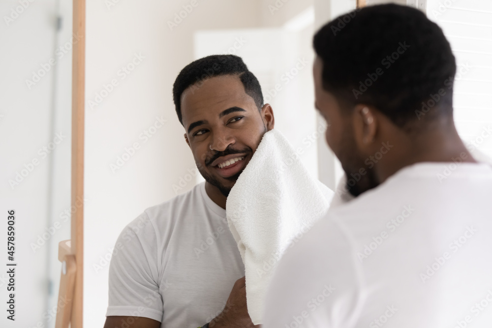 African man in white t-shirt stand in bathroom look in mirror wipes his face with towel after shave, skin care facial procedure smiles feels satisfied. Male beauty cosmetics, skincare, hygiene concept African man in white t-shirt stand in bathroom look in mirror wipes his face with towel after shave, skin care facial procedure smiles feels satisfied. Male beauty cosmetics, skincare, hygiene concept