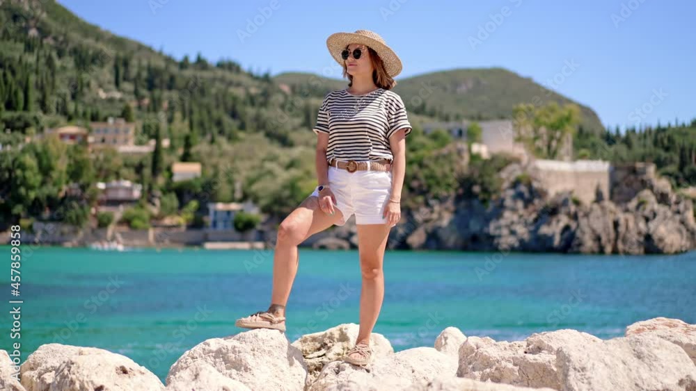 Beautiful warm summer scene - woman in striped t-shirt and hat standing on turquoise sea and mountains background. Girl on stone beach in Greece. Luxury resort concept, traveling